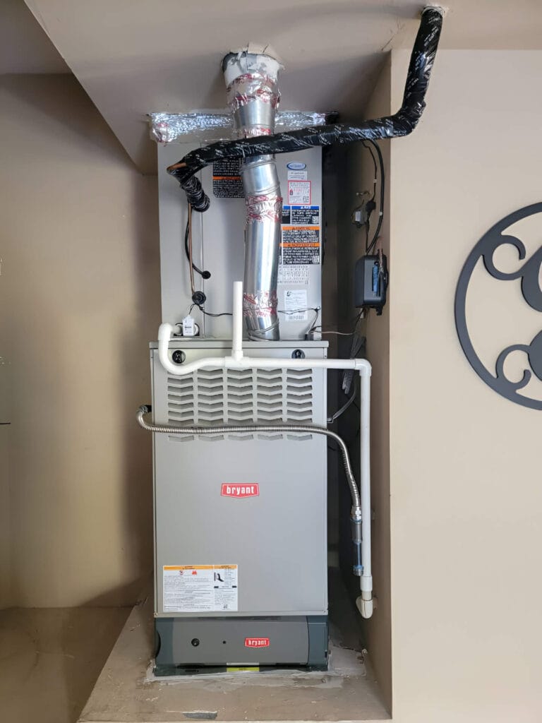 A gray Bryant furnace with ventilation pipes and labels installed in a corner of a room, against a light-colored wall.