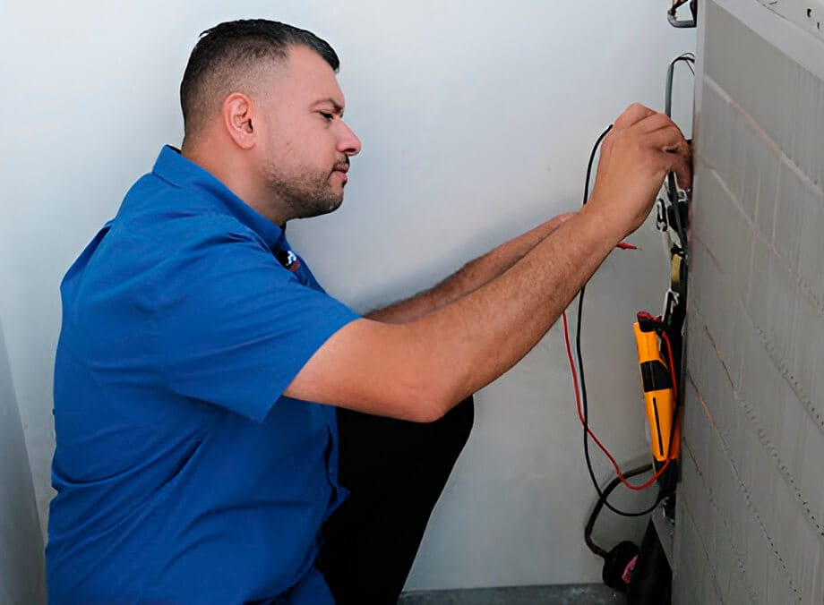 A technician in a blue shirt crouches beside equipment, using tools with wires and a multimeter for electrical diagnosis.