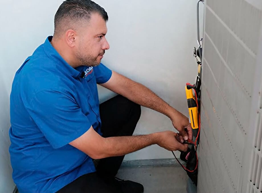 A technician in a blue shirt crouches near an appliance, using tools to perform maintenance or repairs.