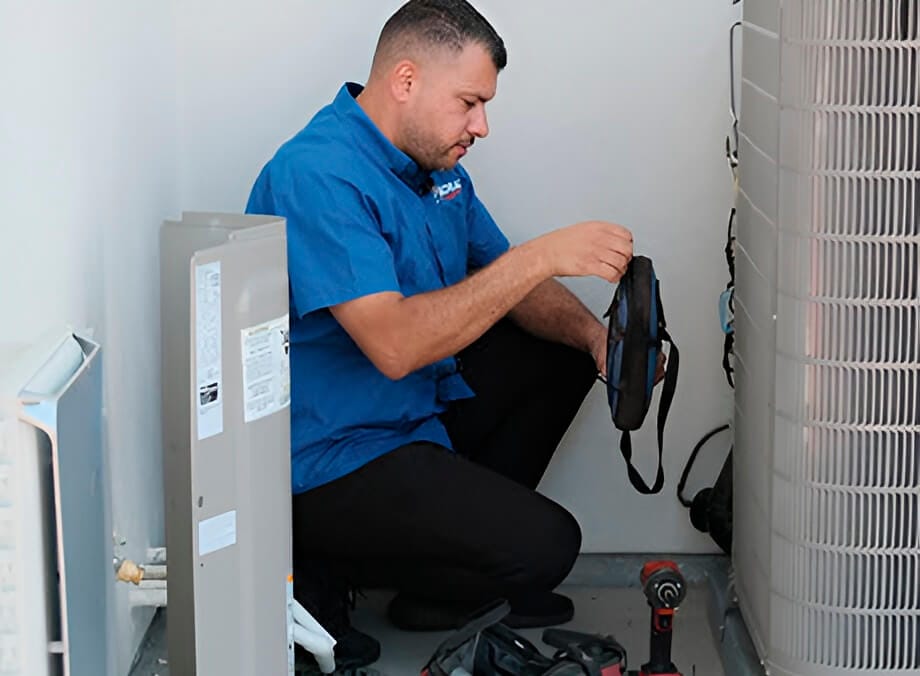 A technician in a blue shirt kneels beside an air conditioning unit, holding a tool bag and preparing to work.
