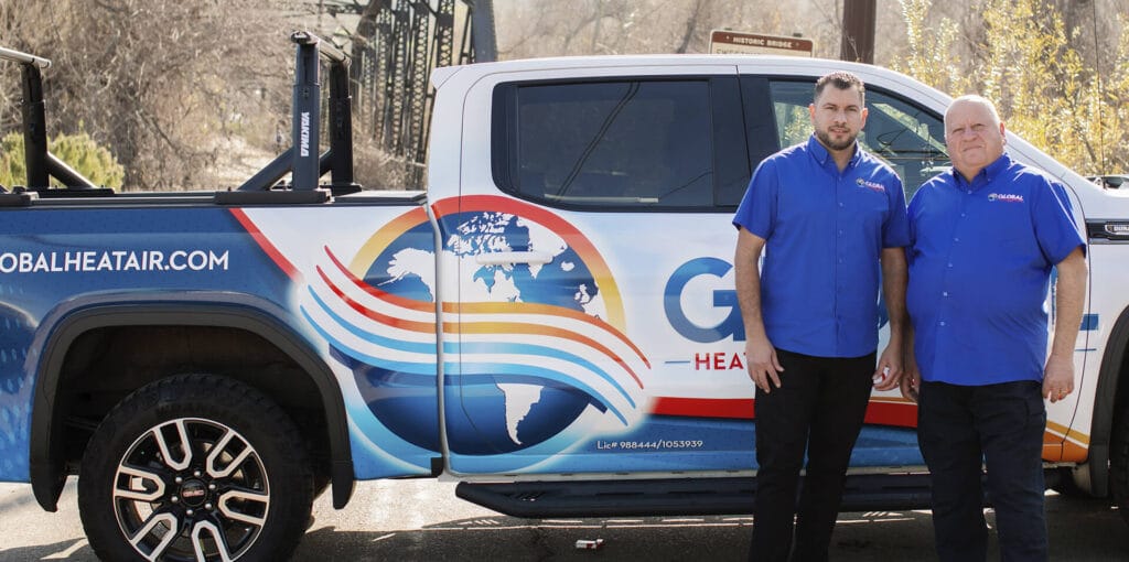 Two men in blue shirts stand beside a branded HVAC service truck.