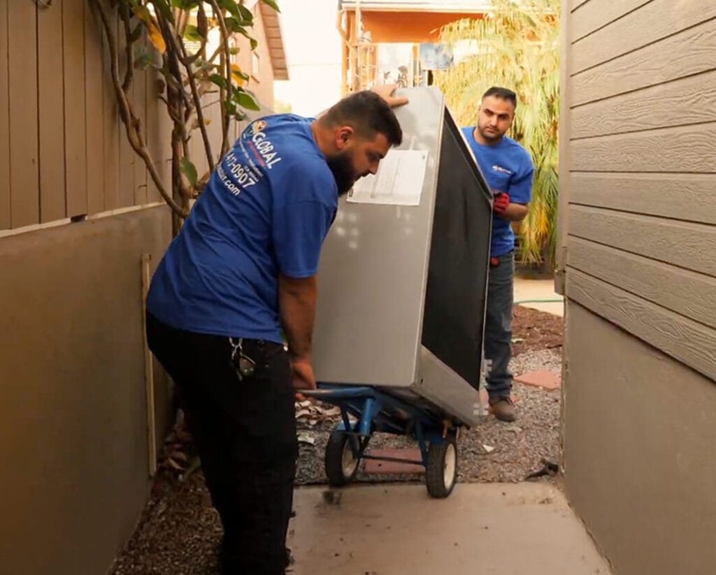 Two workers in blue shirts transport a large appliance.